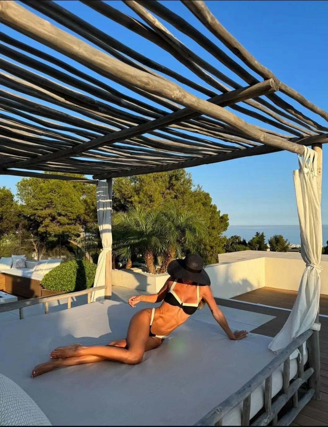 Woman in a black bikini and sun hat lounging on a canopy bed under wooden pergola, enjoying a sunny day with ocean view and trees.