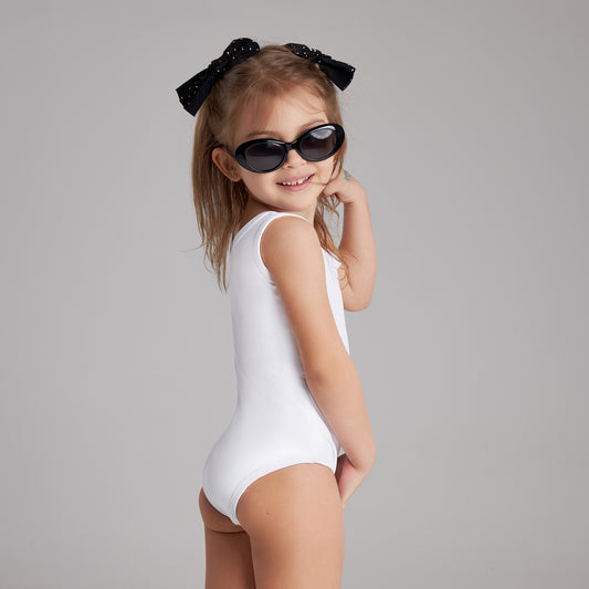 Young girl in a white swimsuit and black sunglasses smiles while posing playfully, wearing a black bow in her hair against a gray backdrop.