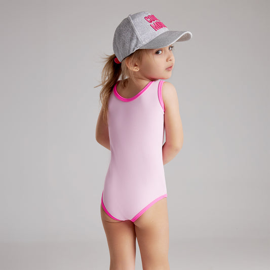 A little girl in a pink swimsuit and a baseball cap smiles playfully at the camera.