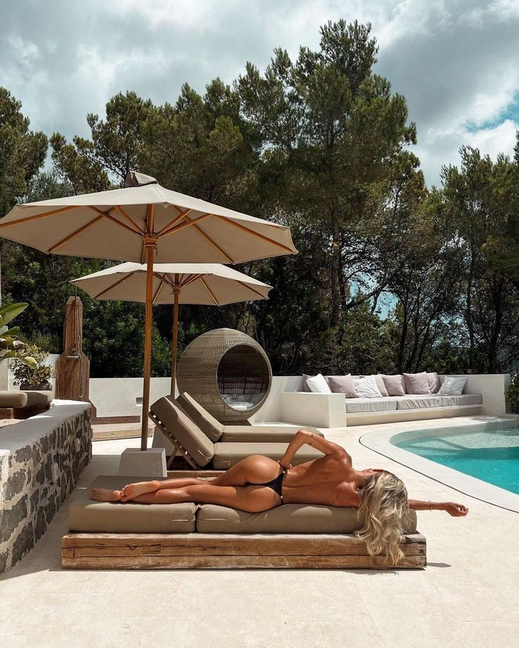 Woman sunbathing on a poolside lounge chair in a tropical resort setting with umbrellas, trees, and outdoor seating in the background.
