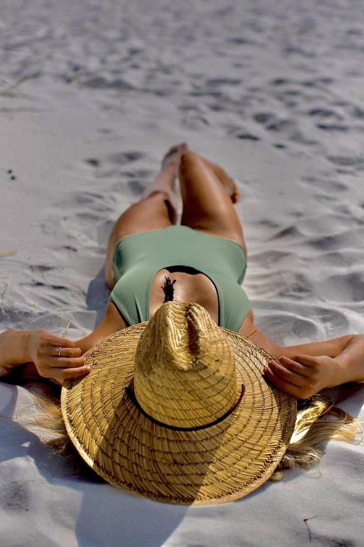 Woman lying on sandy beach wearing a green swimsuit and wide-brim straw hat, relaxing under the sun with her hands behind her head.
