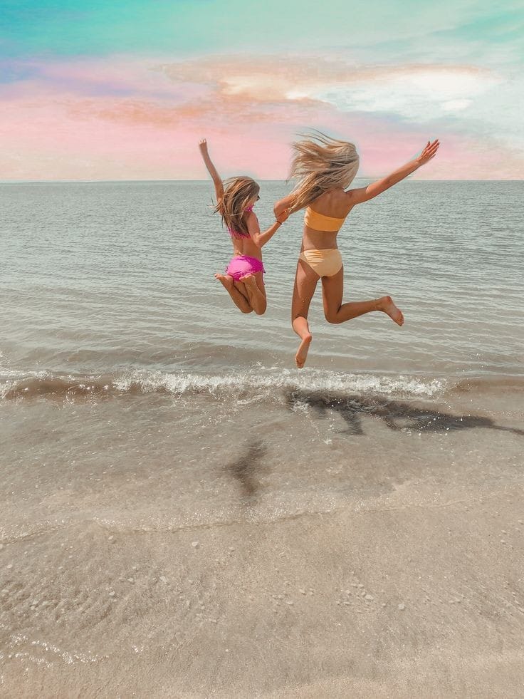 Woman and young girl in bikinis joyfully jumping by the ocean shore under a pastel sky, holding hands in mid-air.