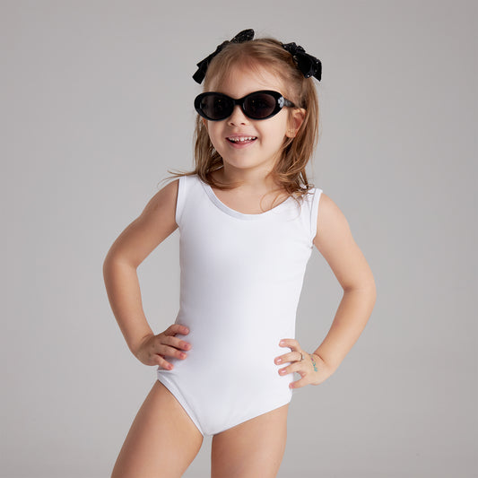 Smiling young girl in a white swimsuit and black sunglasses poses confidently with hands on hips against a plain gray background.